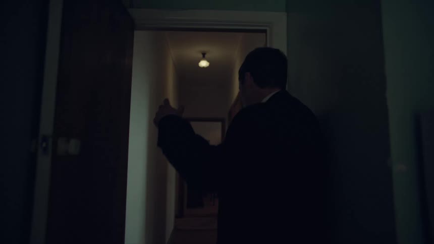 A man in a suit slowly walks down a dim, narrow hallway in a dramatic tracking shot