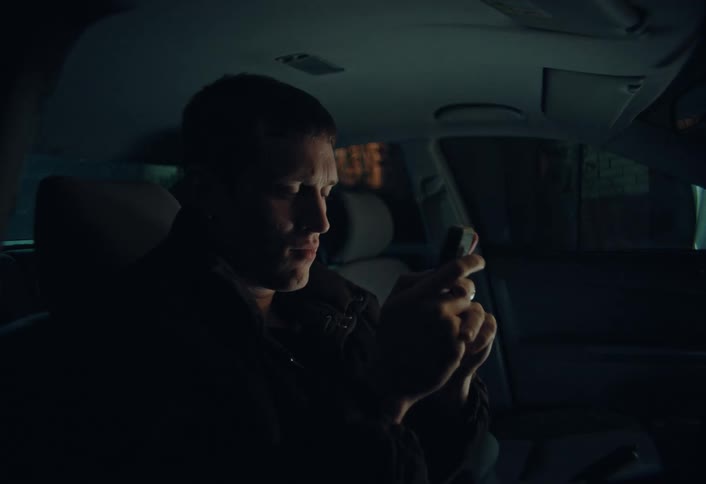 A young man sits in the driver's seat of his car at night, smiling while texting on his smartphone