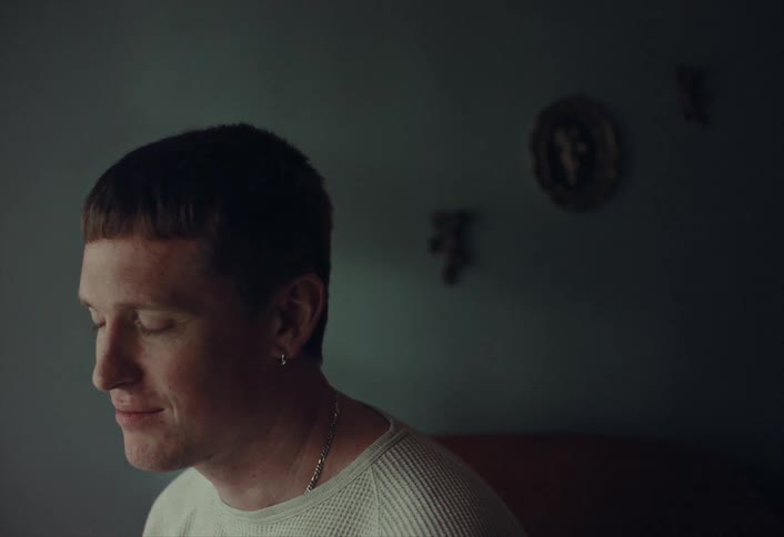 A pensive young man with a modern hairstyle and earring smiles while talking in a softly lit, moody room