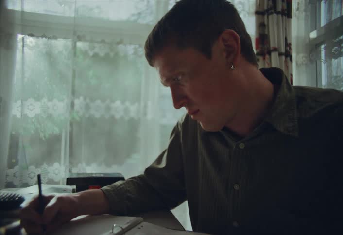 A focused young man studies at a cluttered desk, diligently writing in a notebook by a lace-curtained window