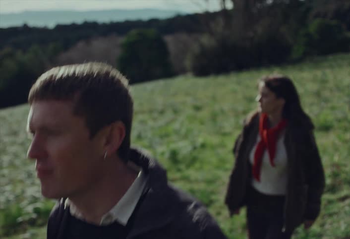 Young man hikes determinedly up a grassy hill in the countryside with a woman following behind him