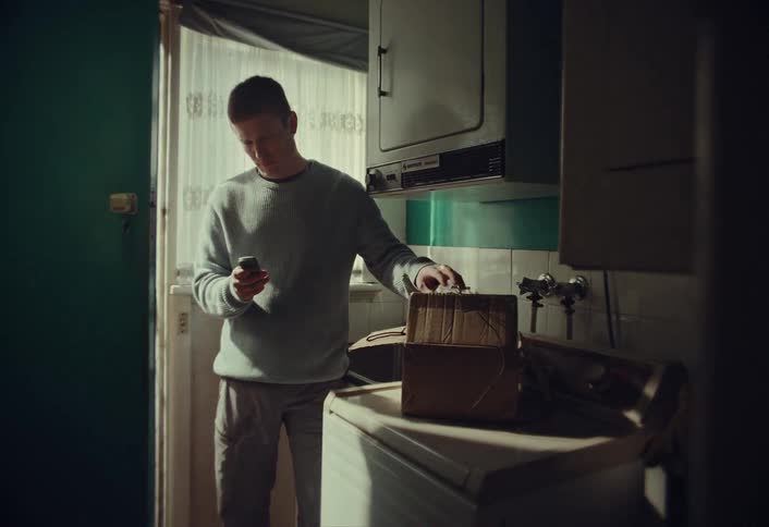 A man in a dimly lit kitchen unpacks and prepares a homemade bomb using his phone