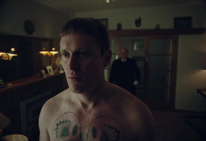 A tense scene unfolds as a butler with a rope approaches a shirtless, tattooed young man from behind in a dimly lit room