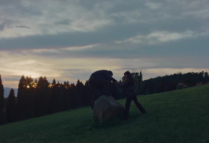 A man and woman hike on a grassy hill at sunset, where the man climbs onto a large rock to take in the view
