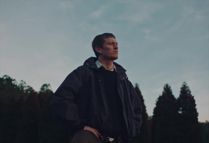 Low-angle shot of a young man in a jacket standing outdoors at dusk, looking thoughtfully into the distance