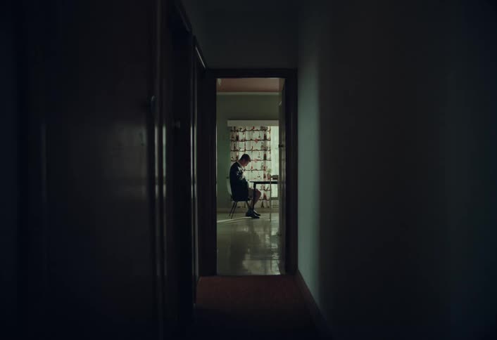 A man sitting at a table in shorts puts on a suit jacket and walks purposefully down a dark hallway toward the camera