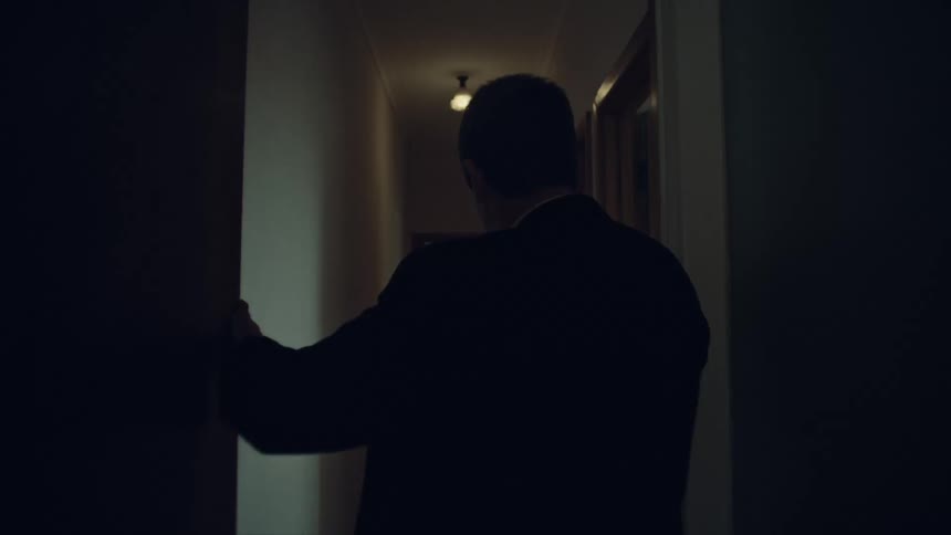 A disoriented man in a suit stumbles drunkenly down a dimly lit hallway, supporting himself against the wall
