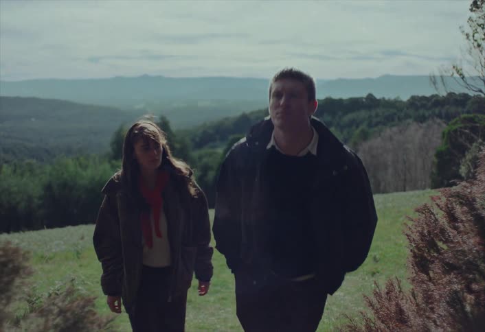 A young man and woman walk uphill together through a scenic, grassy landscape on a cool, overcast day