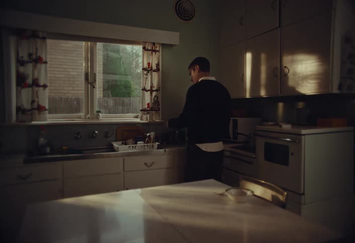 A man stands alone in a dim, retro-style kitchen, opening a bottle of wine as sunlight streams through the window