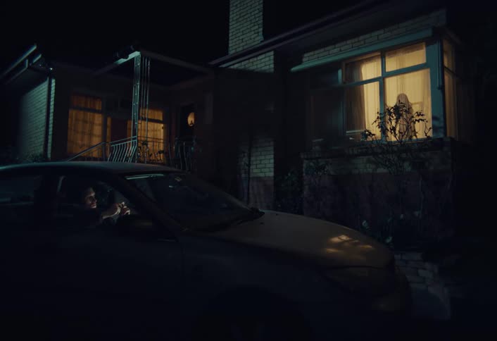 A person sits in their car at night watching a mysterious figure appear in the window of a suburban home