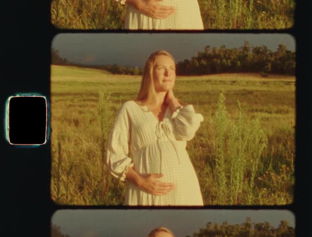 A pregnant woman in a white dress gently holds her belly while standing in a sunlit field, shot on vintage film