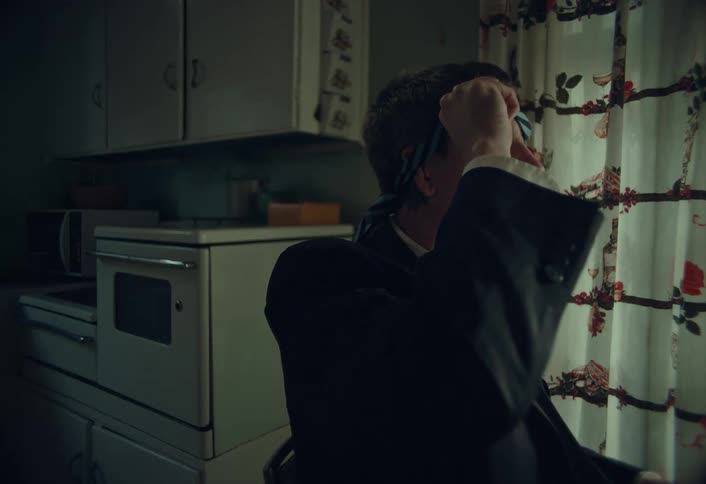 A man in a suit sits in a retro kitchen and slowly takes off his tie blindfold