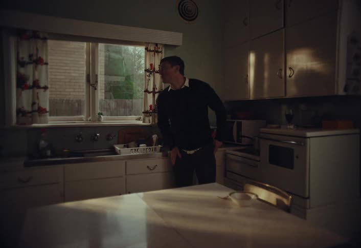 A pensive man stands alone in a dimly lit, retro-style kitchen, looking out the window with a melancholic expression