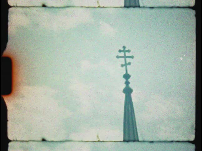 A grainy, vintage 8mm film captures the silhouette of an Orthodox cross atop a church steeple against a cloudy sky
