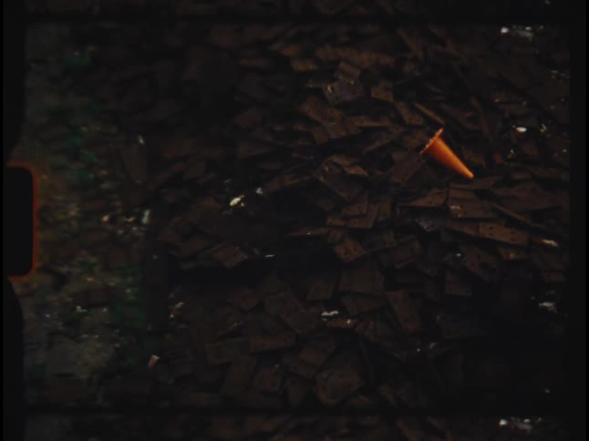 A vintage 8mm film shot zooms out from an orange traffic cone on a pile of scrap metal next to railroad tracks
