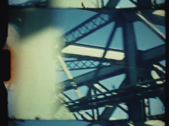 A vintage 8mm film captures an abstract, low-angle view while moving through the steel trusses of a bridge