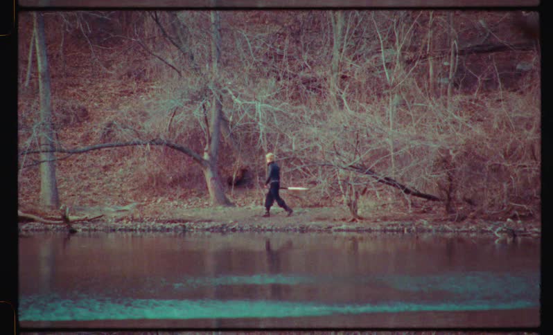 A lone warrior carrying a sword walks along the bank of a lake in a desolate autumn forest