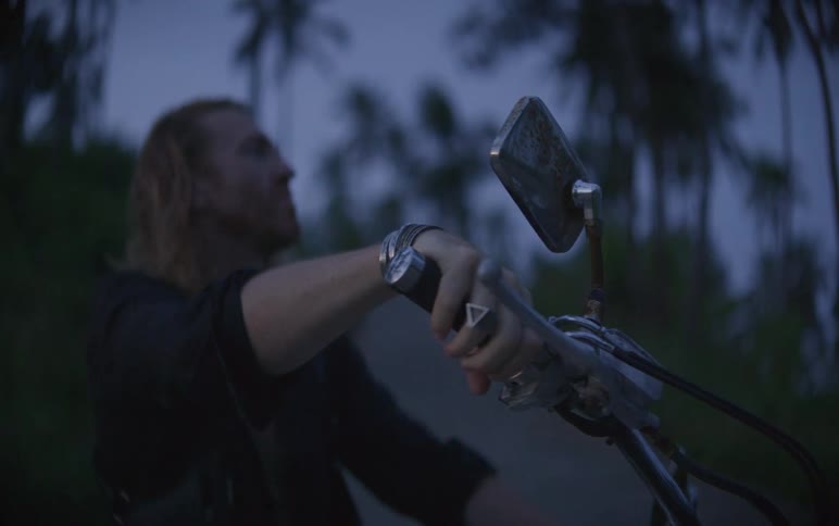A man with long hair revs the throttle of his motorcycle at dusk and then dismounts the bike on a rural road