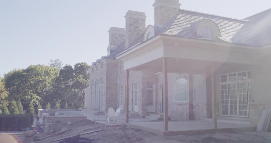 A slow pull-back reveals a luxury stone mansion with a new patio and yard under construction on a sunny day