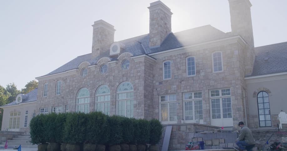 A worker performs landscaping and masonry work in the sun-drenched backyard of a luxurious stone mansion