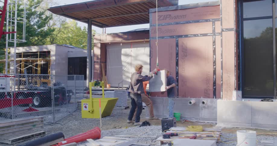 Slow zoom out of three construction workers carefully installing a heavy stone panel on the exterior of a modern building