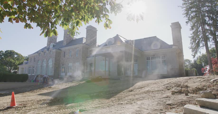 Backlit dolly shot reveals a luxury stone mansion under construction as workers landscape the grounds on a sunny day