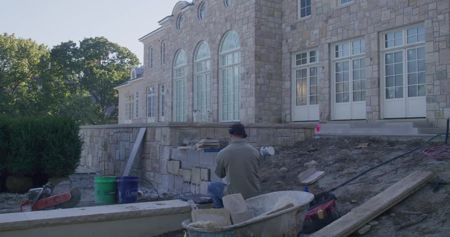 A stonemason carefully constructs a stone retaining wall at a luxury residential construction site on a sunny day