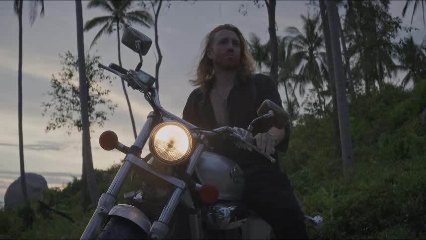 A long-haired man on a chopper motorcycle turns off the ignition and dismounts in a tropical setting at dusk