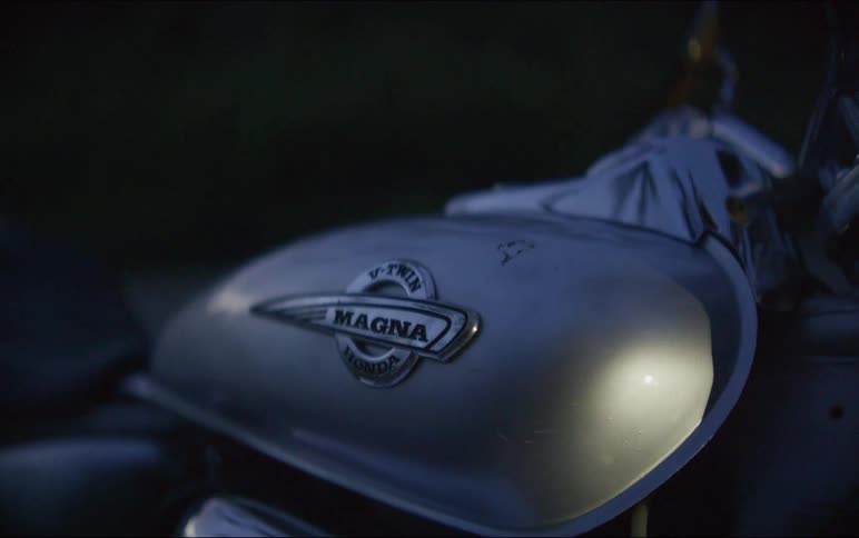 A cinematic close-up pans across a Honda Magna motorcycle's gas tank and handlebars as a beam of light passes over it at dusk