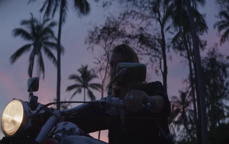 A long-haired biker starts his motorcycle and adjusts his mirrors, silhouetted against a beautiful tropical sunset with palm trees