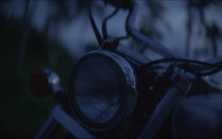 A moody close-up shot of a classic motorcycle's headlight and handlebars parked outdoors at dusk