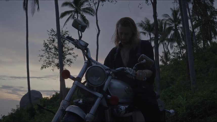 A long-haired biker gets on his chopper motorcycle at dusk with palm trees silhouetted against the evening sky