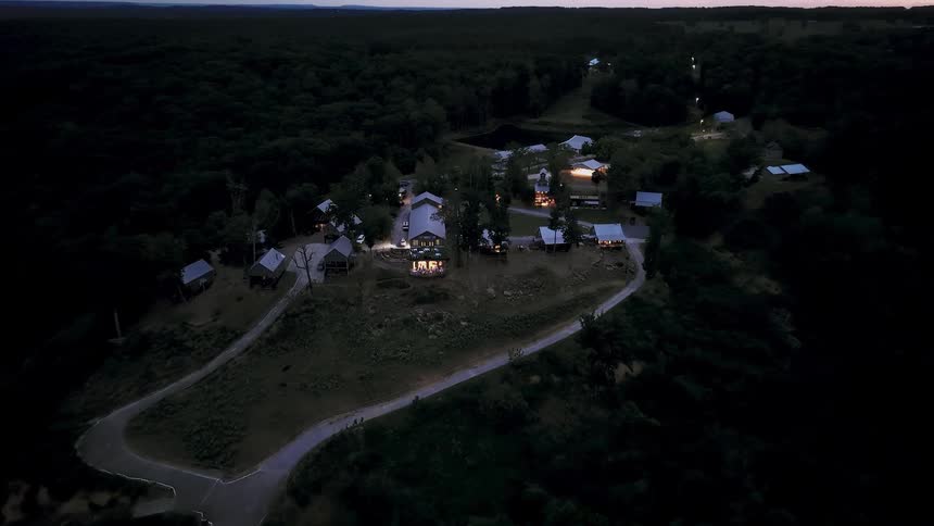 Slow aerial push-in towards a remote, illuminated community of cabins nestled within a dense forest at dusk