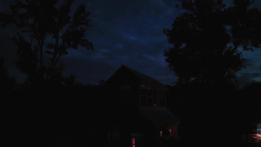 Aerial reveal of a modern cabin at night with colorful party lights inside and a bonfire glowing nearby
