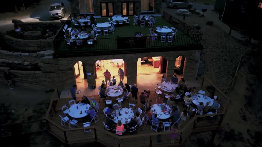 Aerial view rises over a large family gathering enjoying a festive dinner party on multiple outdoor decks at dusk