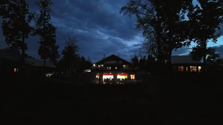 People gather on the brightly lit porch of a rustic lodge as evening falls under a dramatic cloudy sky
