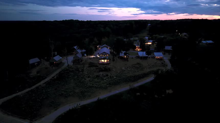 An aerial timelapse reveals a secluded woodland retreat with cozy, lit cabins as the sun sets and twilight fades into night