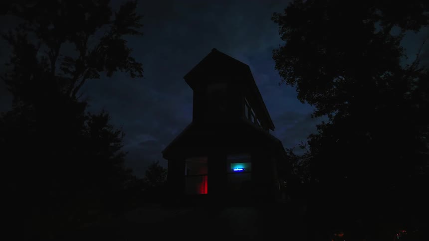 A slow zoom-in time-lapse of a mysterious silhouetted house with colorful glowing windows as the dark night sky transitions to a purple dawn