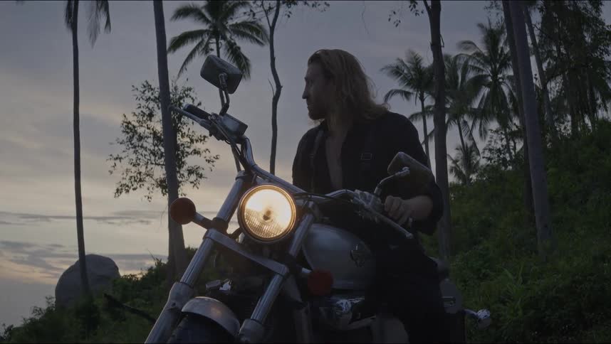 A long-haired man in black sits on a motorcycle with the headlight on at dusk, then turns it off and dismounts in a tropical setting with palm trees