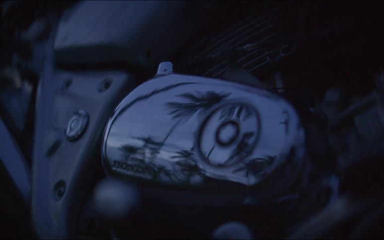 A moody close-up of a Honda motorcycle engine cover reflects palm trees silhouetted against a dark blue evening sky
