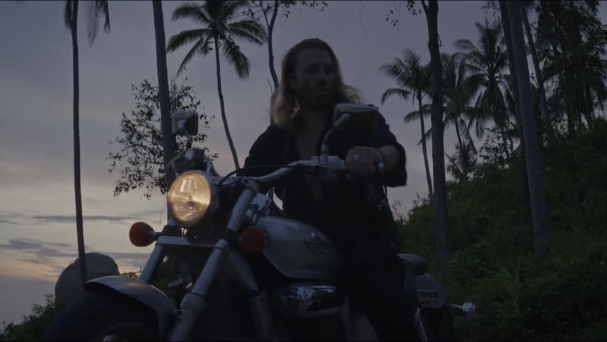 A long-haired biker gets on his chopper motorcycle at dusk and starts the engine with tropical palm trees in the background