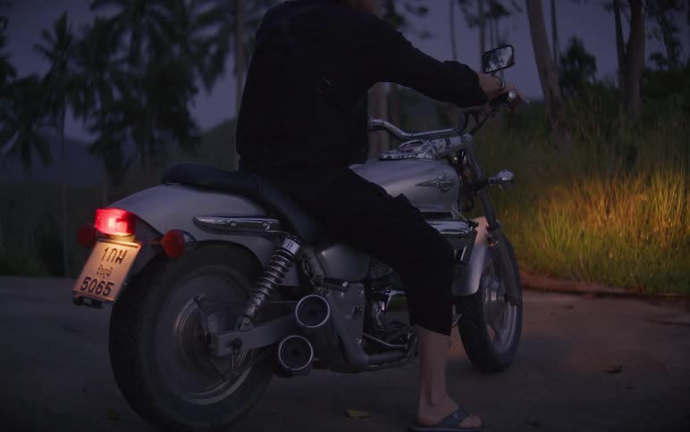 A person in black gets onto a silver motorcycle, turns on the headlights, and slowly drives away down a road at dusk