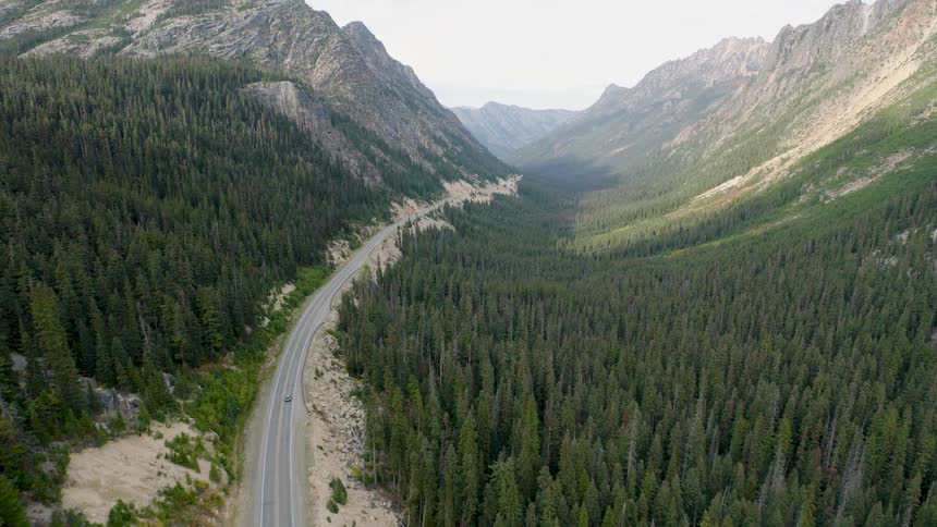 Aerial footage follows a car driving on a scenic highway through a vast mountain valley surrounded by a dense evergreen forest