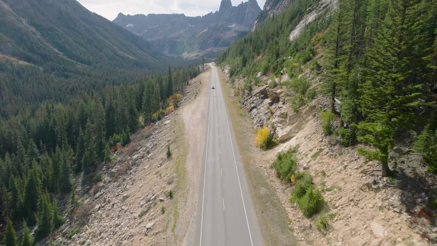An aerial view follows a white SUV driving down a scenic highway through a vast mountain valley