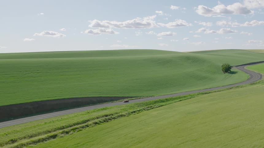 An aerial view of a red convertible sports car driving along a winding road through beautiful, vast rolling green hills on a sunny day