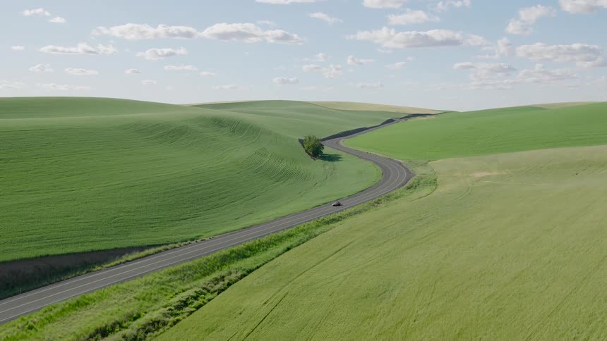 Aerial view of a red car driving on a scenic, winding country road through vast, rolling green hills under a partly cloudy sky