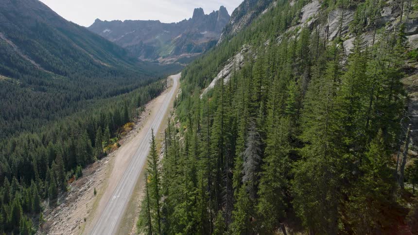 An aerial view reveals a lone motorcyclist riding down a remote mountain pass surrounded by a vast evergreen forest and towering peaks