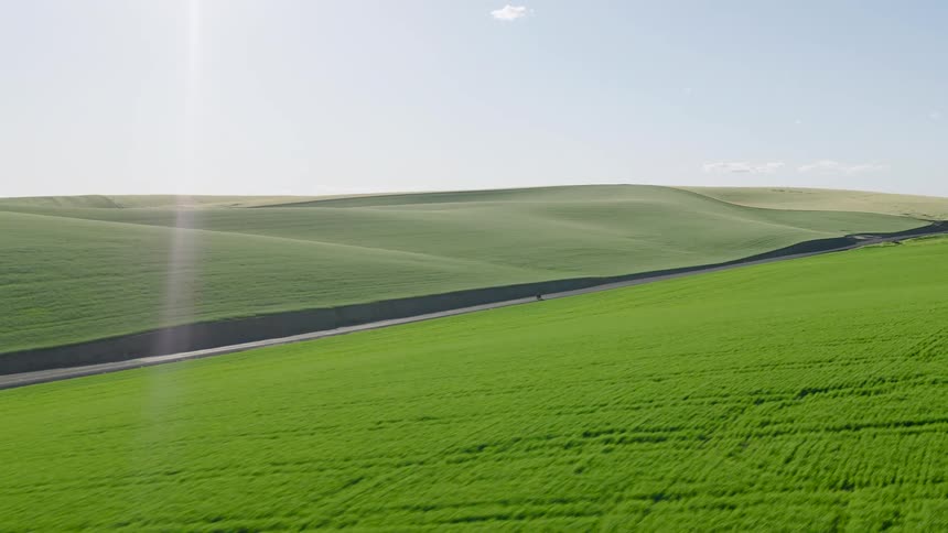 A lone cyclist rides down a country road through vast, rolling green hills on a sunny day
