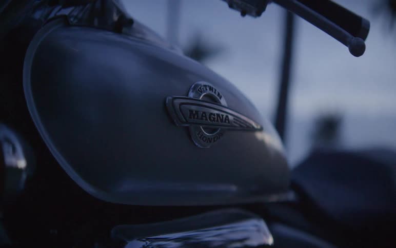 A moody close-up shot focuses on the chrome emblem of a Honda Magna motorcycle at twilight