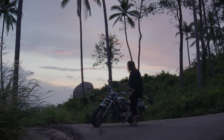 A long-haired biker walks to their motorcycle parked on a tropical hillside road and prepares to ride off into the dusk
